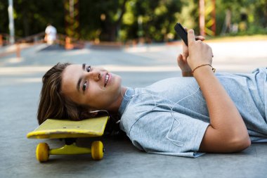 Genç adam paten parkta vakit cep telefonu tutan kaykay üzerinde döşeme kulaklık ile müzik dinlemek gülümseyen