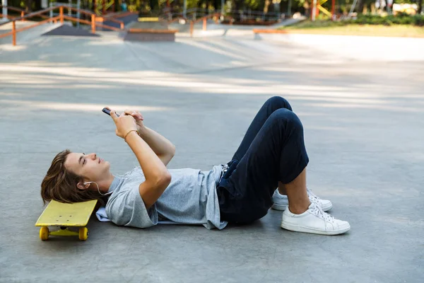 携帯電話を保持しているスケート ボードの上に敷設 ヘッドフォンで音楽を聴くスケート公園で時間を過ごす若い男の笑みを浮かべてください — ストック写真