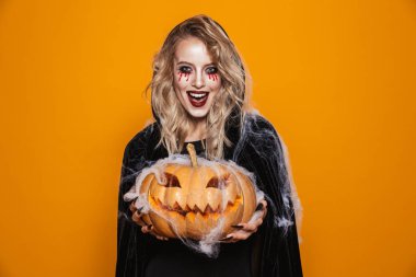 Kabak tutarak ve turuncu üzerinde izole kamera için gülüyor siyah pelerin içinde mutlu korkunç sarı cadı