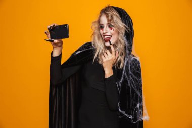 Güzel sarışın Bayan cadı selfie smartphone yapma ve turuncu üzerinde izole gülümseyen korkunç makyaj ile