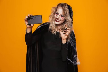 Güzel sarışın Bayan cadı selfie smartphone yapma ve turuncu üzerinde izole gülümseyen korkunç makyaj ile