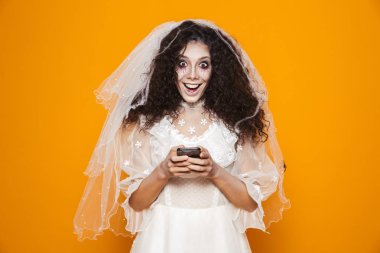 Beyaz elbiseli mutlu ölü gelin kamera seyir ve smartphone kullanarak bir peçe üzerinde turuncu izole.