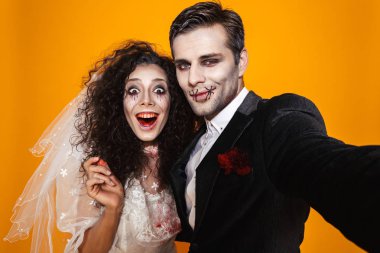 Fotoğraf eğlenceli zombi çift damat ve gelin sarı arka plan üzerinde izole selfie çekerken gülüyor düğün kıyafeti ve Cadılar Bayramı makyaj
