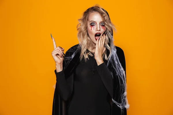 Shoked woman witch with opened mouth wearing black dress and cloak and pointing up at copy space isolated over orange