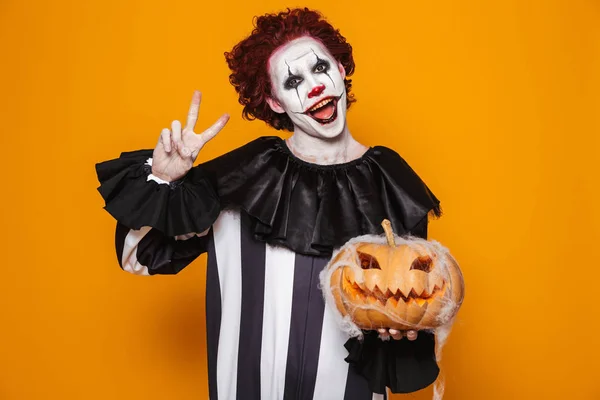 Clown happy man wearing black costume and halloween makeup looking at camera isolated over yellow background showing peace gesture holding pumpkin.