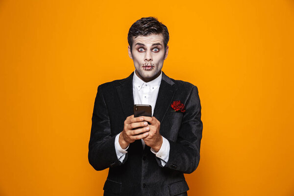 Photo of young dead gentleman on halloween wearing classical suit and creepy makeup holding mobile phone isolated over yellow background