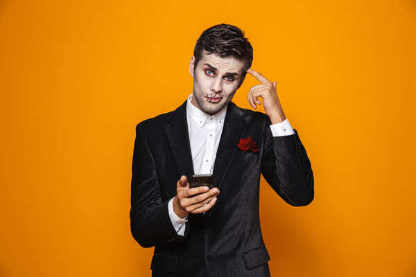 Photo of handsome dead man on halloween wearing classical suit and creepy makeup holding mobile phone isolated over yellow background