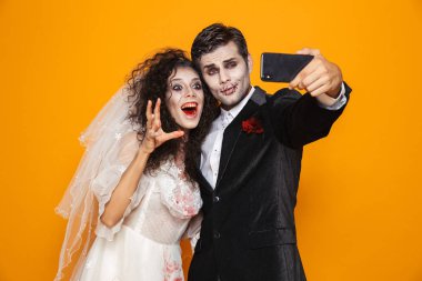 Neşeli zombi çift damat ve gelin alma selfie sarı arka plan üzerinde izole ederken gülen düğün kıyafeti ve Cadılar Bayramı makyaj fotoğrafı