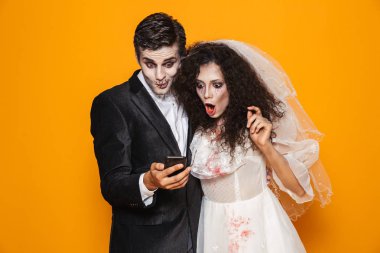 Şaşırmış ürpertici adam ve kadın Cadılar Bayramı kostümleri üzerinde turuncu izole smartphone kullanarak düğün