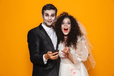 Şaşırmış mutlu ölü gelin ve damat kamera seyir ve smartphone üzerinde turuncu izole tutan hallowen kostümleri