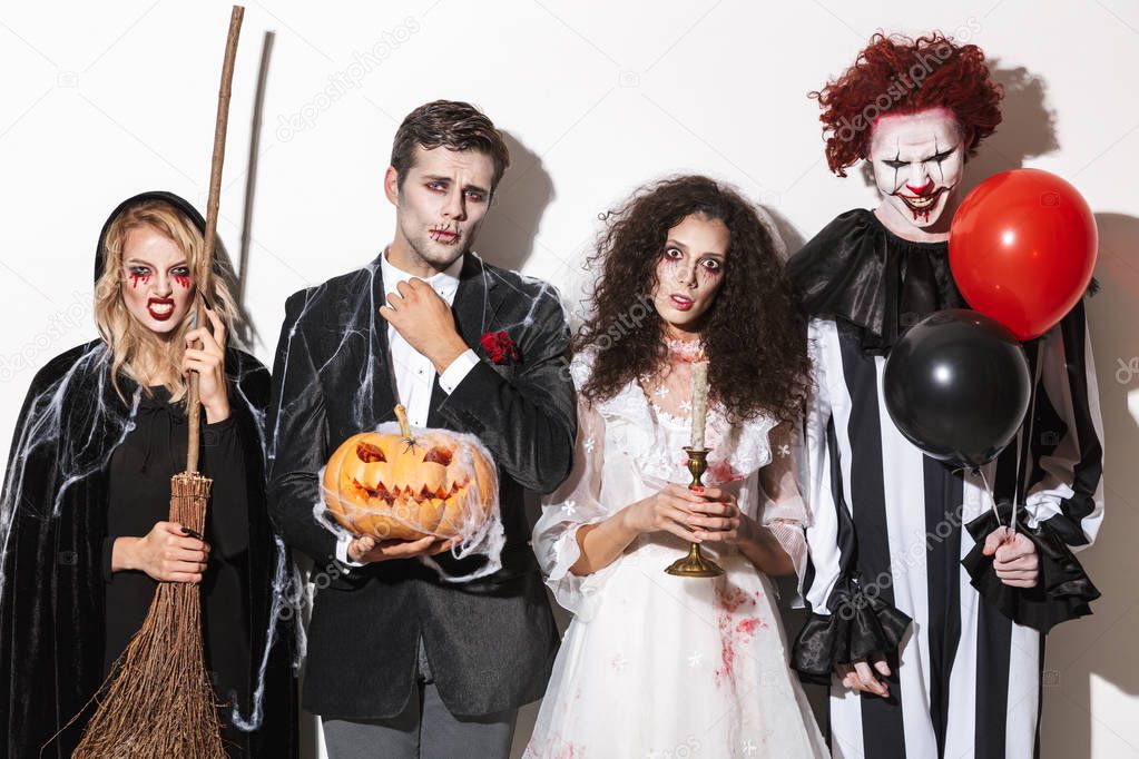 Group of friends dressed in scary costumes celebrating Halloween isolated over white background, holding balloons, curved pumpkin