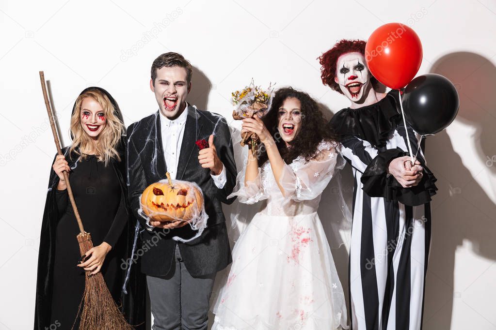 Group of joyful friends dressed in scary costumes celebrating Halloween isolated over white background, holding balloons, curved pumpkin