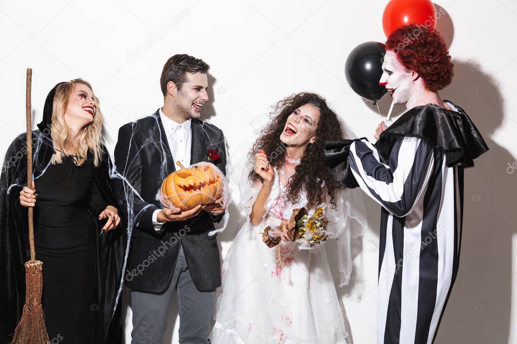 Group of happy friends dressed in scary costumes celebrating Halloween isolated over white background, holding balloons, curved pumpkin, broom