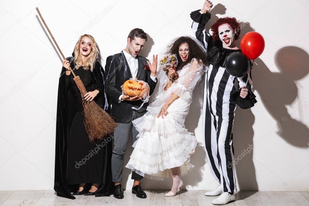 Group of cheerful friends dressed in scary costumes celebrating Halloween isolated over white background, holding balloons, curved pumpkin, broom