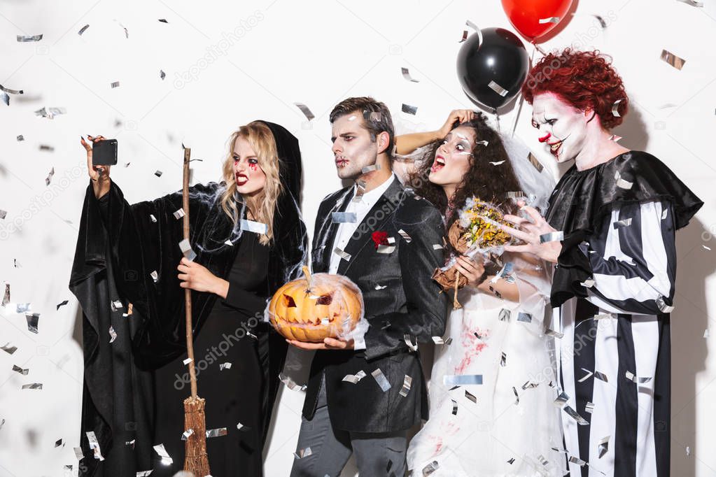 Group of excited friends dressed in scary costumes celebrating Halloween under confetti rain isolated over white background, holding balloons and curved pumpkin, taking a selfie