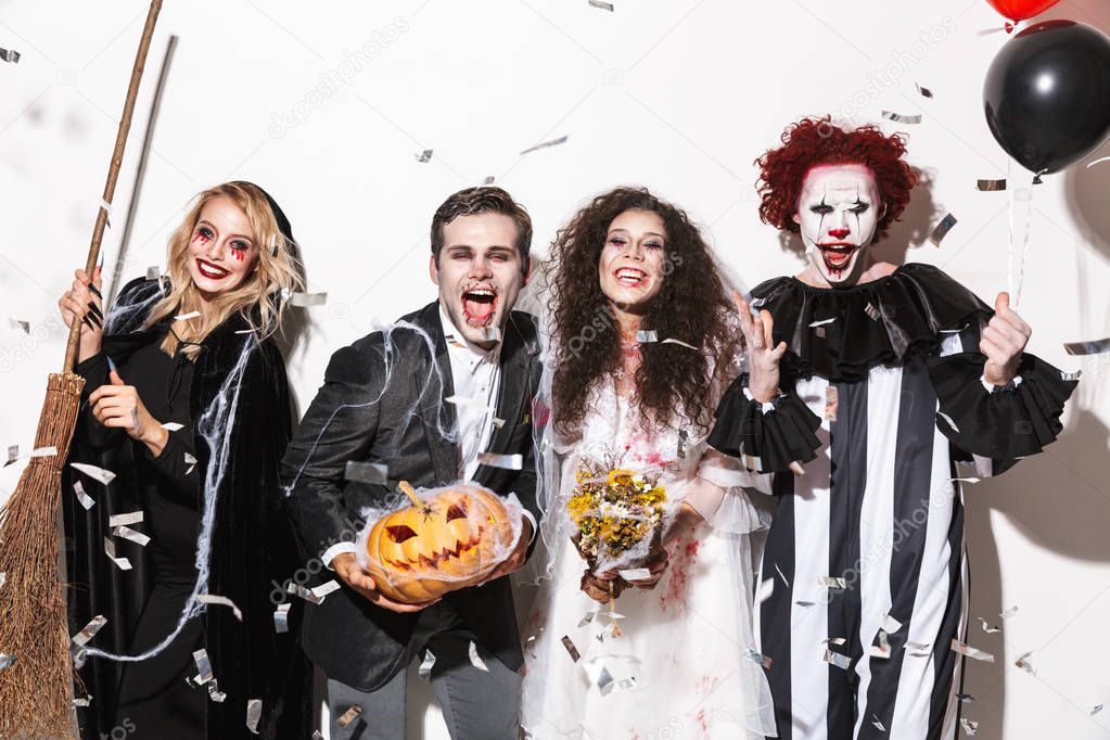 Group of joyful friends dressed in scary costumes celebrating Halloween under confetti rain isolated over white background, holding balloons, curved pumpkin, broom