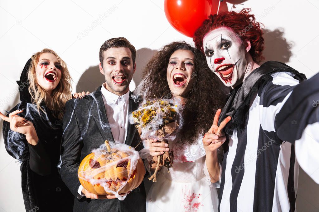 Group of excited friends dressed in scary costumes celebrating Halloween under confetti rain isolated over white background, holding balloons and curved pumpkin, taking a selfie