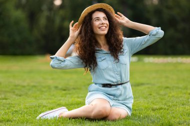 Hasır şapka ve çim parkta oturan elbise giyen uzun siyah saçlı güzel esmer kadın görüntüsünü