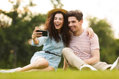 Görüntü çift erkek ve kadın 20'li yaşlarda park ve alma selfie Smartphone cep telefonu ile yeşil çimenlerin üzerinde oturan