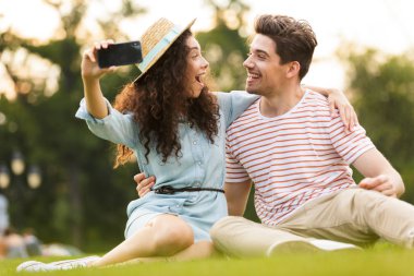 Eğlenceli çift insan görüntüsünü ve kadın 20'li yaşlarda oturan park ve cep telefonu alarak selfie yeşil çimenlerin üzerinde