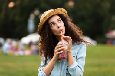 Görüntü gülümseyen ve arka plan bulanık ile yeşil parkta yürürken plastik bardak içecek içme kafkas kadın 20'li yaşlarda giyen hasır şapka