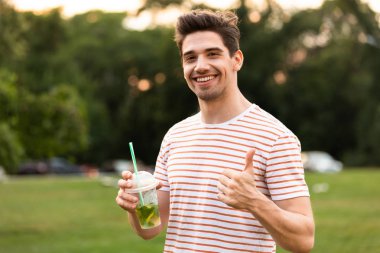 Görüntü gülümseyen adam parkta yürüyüş ve plastik bardak içecek içme 20s
