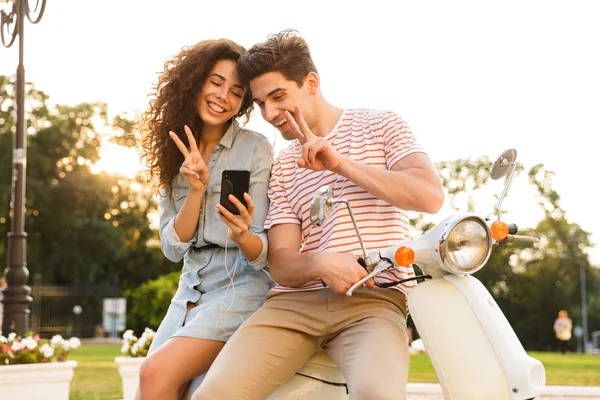 美丽的夫妇的肖像 穿着耳机在智能手机微笑 而坐在摩托车一起在城市街道 — 图库照片