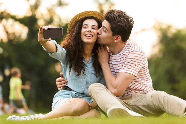 美しいカップル男性と女性 代の画像公園で携帯電話で撮影 Selfie 緑の草の上に座って — ストック写真
