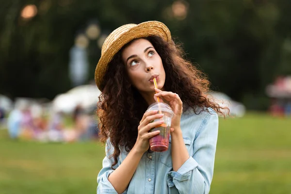 Görüntü gülümseyen ve arka plan bulanık ile yeşil parkta yürürken plastik bardak içecek içme kafkas kadın 20'li yaşlarda giyen hasır şapka