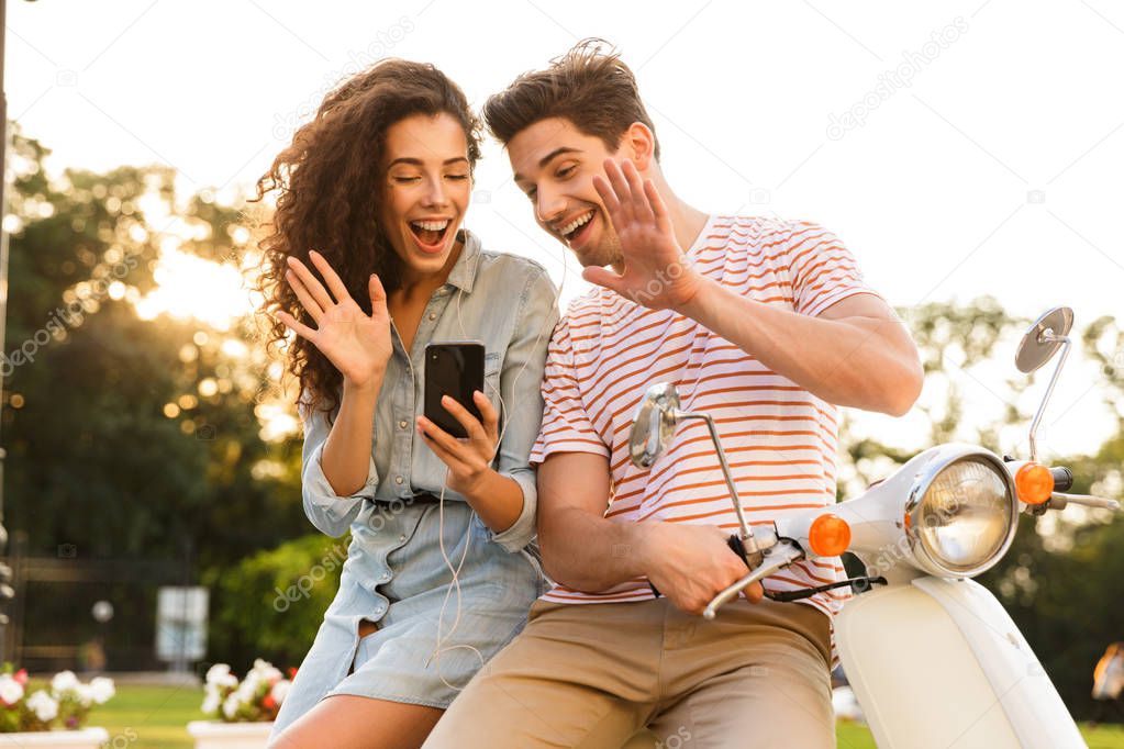 Portrait of happy couple man and woman wearing earphones looking at smartphone while sitting on scooter together on city street