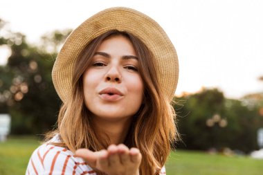 Bir öpücük gönderme gülümseyen genç kız parkta, çimlerin üzerine oturan yaz şapkalı yakın çekim