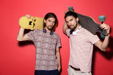 İki genç çekici ikiz kardeşler portresi kulaklık ile müzik dinlemek, kaykaylar tutan kırmızı arka plan üzerinde izole