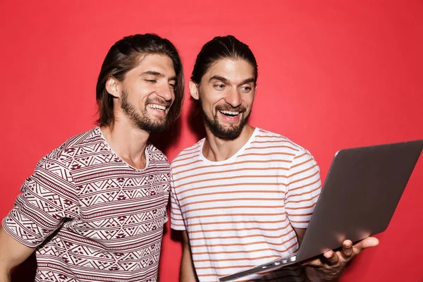 Portrait of a two young smiling twin brothers isolated over red background, holding laptop computer