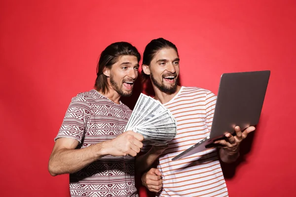 Portrait of a two young happy twin brothers isolated over red background, holding laptop computer, showing money banknotes