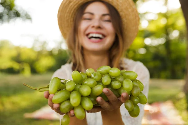 关闭笑年轻的女孩在夏天的帽子花时间在公园里 显示绿色的葡萄 — 图库照片