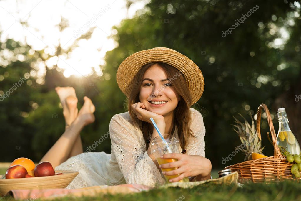 Lovely young girl in summer hat having a picnic at the park, laying on a grass, drinking juice