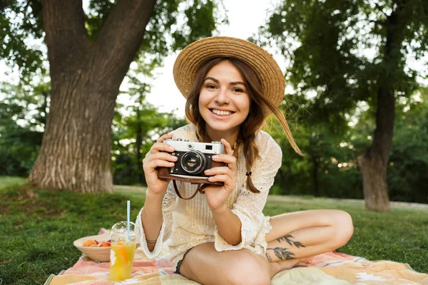 微笑的年轻女孩在夏天的帽子在公园野餐 坐在草地上 拿着照片相机 — 图库照片