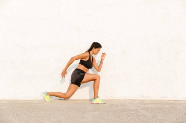 Tam boy görüntü esmer kızın 20'li yaşlarda çömelme ve duvardan vücudu germe eşofman