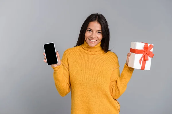 一个微笑的休闲年轻女子的肖像孤立在灰色背景 显示空白屏幕手机 拿着礼物盒 — 图库照片