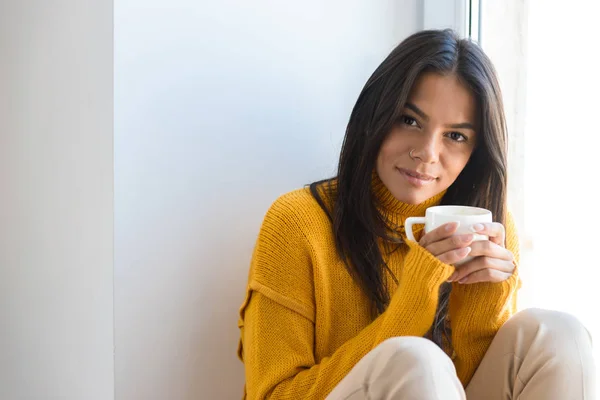 素敵な若い女性の室内では 窓際に座ってセーターに身を包んだお茶のカップを保持している肖像画間近します — ストック写真