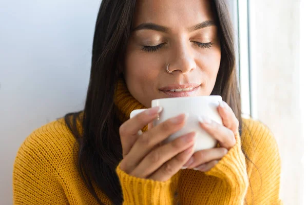 素敵な若い女性の室内では 窓際に座ってセーターに身を包んだお茶を飲んで肖像画間近します — ストック写真