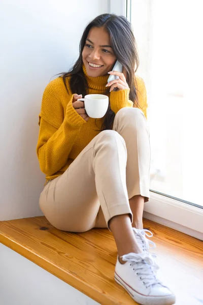 飲むコーヒーが電話で話している灰色の壁背景に分離された窓の近くに座っている幸せな女性のイメージ — ストック写真