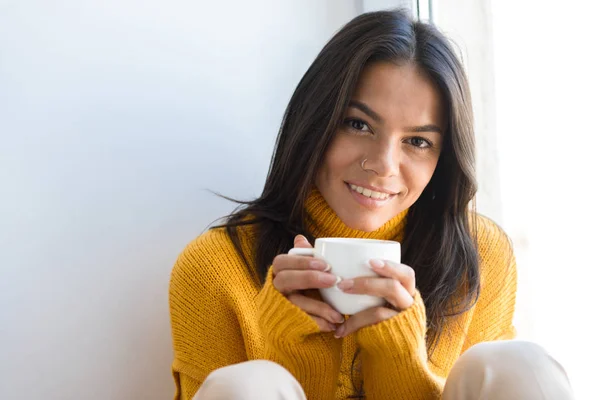 笑顔の若い女性の室内では 窓際に座ってセーターに身を包んだお茶のカップを保持している肖像画間近します — ストック写真