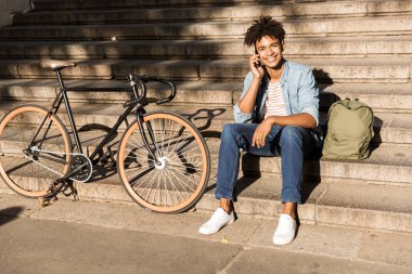 Cep telefonu üzerinde konuşurken ile bisiklet, merdivenlerde oturan neşeli Afrika olan genç adam açık havada, sırt çantası