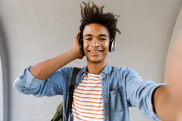 ヘッドフォンで音楽を聴く屋外に立っている陽気な若いアフリカ人のイメージは カメラによる Selfie を作る — ストック写真