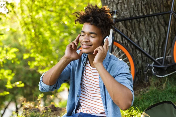 愉快的年轻非洲青少年与背包户外 坐在草地上 听音乐与耳机 — 图库照片