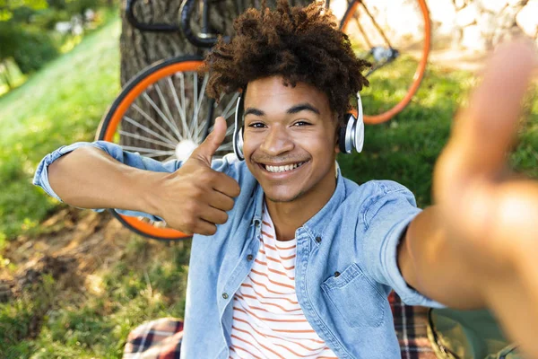 陽気な若いアフリカとティーンエイ ジャー自転車アウトドア 草の上に座って ヘッドフォンで音楽を聴く Selfie 表示を取って親指します — ストック写真