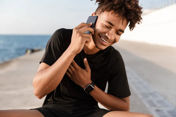 Close up of a young african sportsman at the beach, talking on mobile phone, laughing