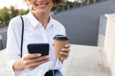 Gülen bir genç kadının sokakta ayakta, kahve holding, cep telefonuyla görüntü kırpılmış