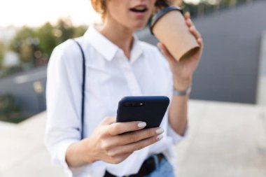 Sürpriz bir genç kadının sokakta ayakta, kahve holding, cep telefonuyla görüntü kırpılmış